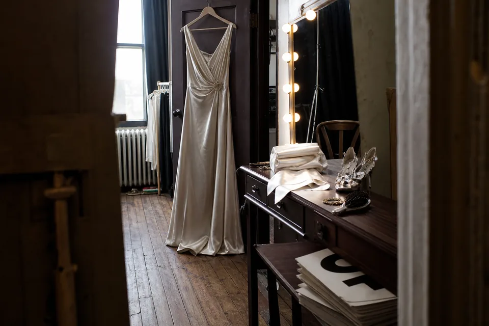 Backstage dressing room in a theater with a silk gown on a wooden hanger, a lit mirror and a folded satin sash in afternoon light