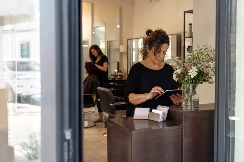 A middle-aged salon owner at the reception desk of her beauty salon with a tablet, business cards and flowers on the counter in morning light