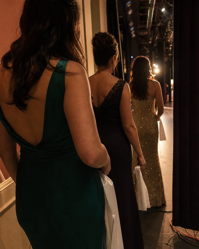 Three women in evening gowns seen from behind at a stage entrance of a hotel ballroom, holding sashes